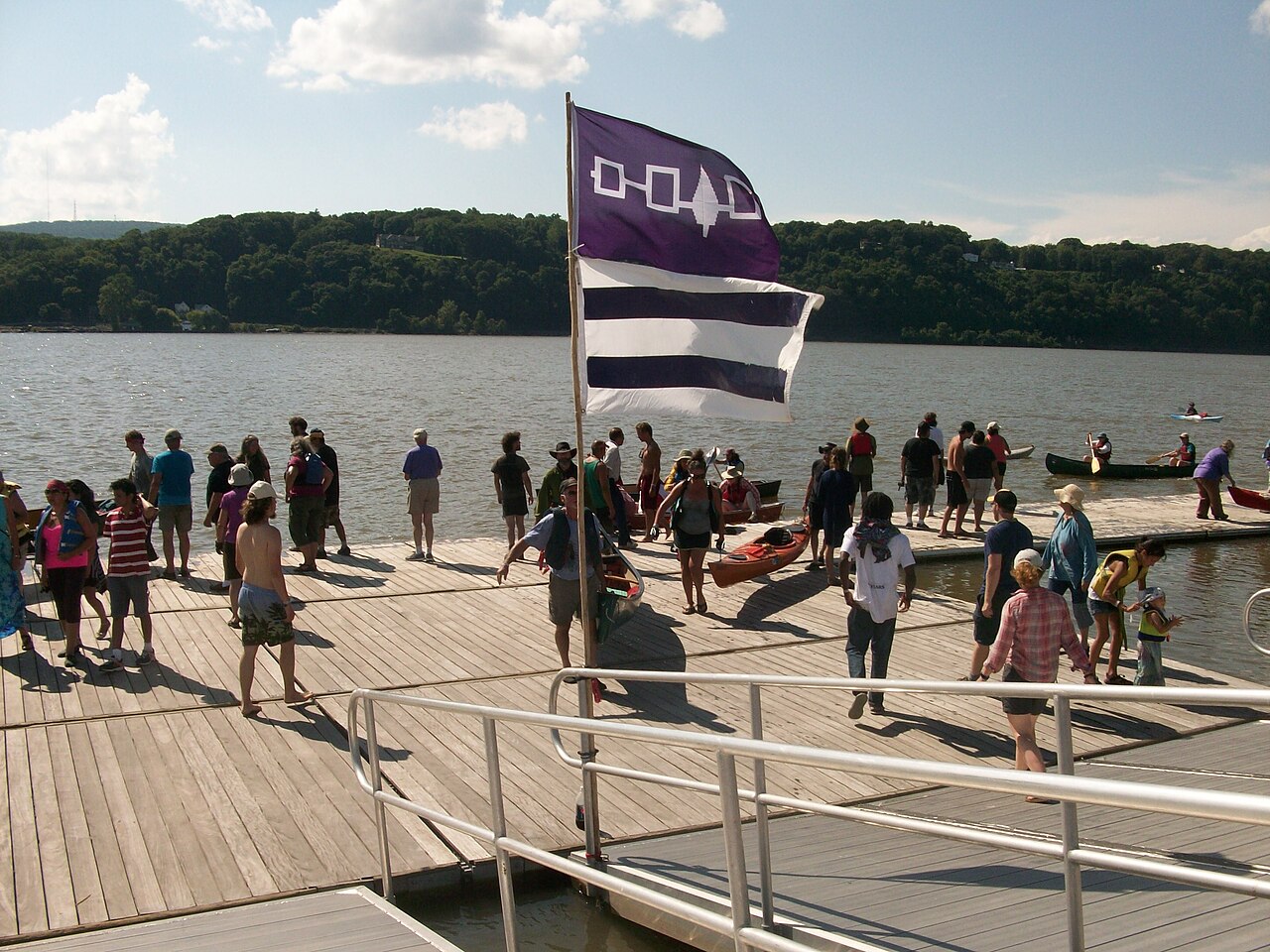 A purple Haudenosaunee flag and a striped Two Row flag fly over paddlers on a river dock